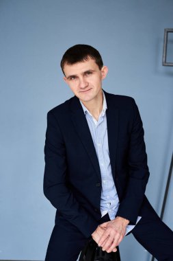 Koyu mavi takım elbiseli, açık mavi gömlekli genç esmer adam, mavi duvarın önünde sinema lambasıyla oturuyor. İş adamı, özgeçmiş için poz veriyor. Şirket kültürü. Duyguların yansıması.