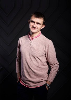 Genç esmer adam, günlük açık pembe kazak giyiyor, fotoğraf stüdyosundaki siyah duvarın önünde poz veriyor, gülümsüyor, gülüyor, elleri ceplerinde. İş adamı şirket kültürü. Mutlu Duygular.