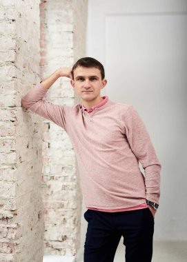 Genç esmer adam, gündelik açık pembe kazak giyiyor, aydınlık odada beyaz tuğla duvara yaslanıyor. Şirket iş kültürü. Duygular. İş adamı portresinin dörtte üçü boş gününde dinleniyor..