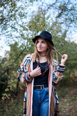 Autumn Park Ormanı 'ndaki güzel sarışın kadının çeyrek portresi. Siyah şapkalı, mavi kot pantolonlu ve doğal arka planda poz veren renkli hırkalı genç bir kız. Hippi tarzı..