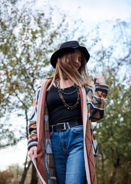 Güz parkı ormanlarında yürüyen sarışın güzel bir kadının dörtte üçü. Siyah şapkalı, mavi kot pantolonlu ve doğal arka planda poz veren renkli hırkalı genç bir kız. Hippi tarzı..