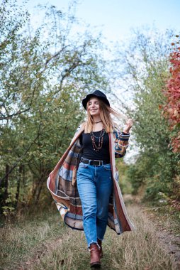 Güz parkı ormanlarında yürüyen sarışın güzel bir kadının dörtte üçü. Siyah şapkalı, mavi kot pantolonlu ve doğal arka planda poz veren renkli hırkalı genç bir kız. Hippi tarzı..