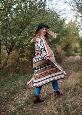 Güz parkı ormanlarında yürüyen sarışın güzel bir kadının uzun bir portresi. Siyah şapkalı, mavi kot pantolonlu ve doğal arka planda poz veren renkli hırkalı genç bir kız. Hippi tarzı..