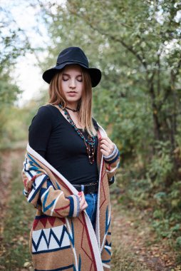 Autumn Park Ormanı 'ndaki güzel sarışın kadının çeyrek portresi. Siyah şapkalı, mavi kot pantolonlu ve doğal arka planda poz veren renkli hırkalı genç bir kız. Hippi tarzı..