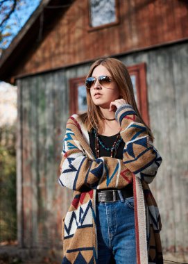 Genç sarışın kadın, renkli kahverengi hırka ve güneş gözlüğü takıyor, sonbaharda köyde eski zamanlardaki ahşap kulübenin önünde poz veriyor. Hafta sonları dışarıdaki güzel hippi kız düşünüyor. Üç çeyrek portre.