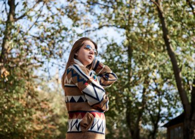 Renkli kahverengi hırkayla kaplanmış, sonbaharda güneşli orman ormanlarında dikilen güzel sarışın kadının dörtte üçlük portresi. Hafta sonu parkta yürüyüş. Hippi tarzı.