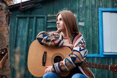 Genç sarışın kadın, renkli hırka giyiyor, akustik gitar tutuyor, sonbaharda ormandaki eski yeşil ahşap kulübenin önündeki bankta oturuyor. Köydeki hippi müzisyenin çeyrek portresi.