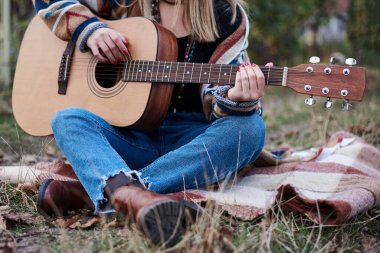 Renkli hırka ve kot pantolon giyen, elinde akustik gitar tutan, sonbaharda kırsal kesimde battaniyede oturan genç sarışın bir kadın. Hippi müzisyenin elindeki gitarın yakın plan fotoğrafı.
