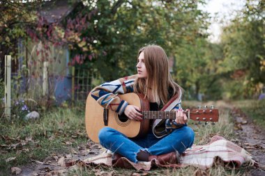 Renkli hırka ve kot pantolon giyen, elinde akustik gitar tutan, sonbaharda kırsal kesimde battaniyede oturan genç sarışın bir kadın. Hippi müzisyen, köy parkında gitar çalıyor.