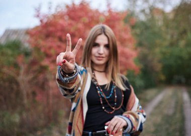 Akustik gitarlı hippi müzisyen, iki parmağıyla işaret veriyor. Renkli hırka ve kot pantolon giyen, sonbaharda orman parkında poz veren genç bir kadının dörtte üçü..