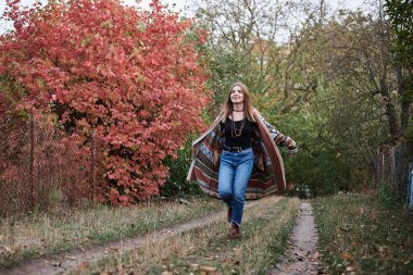 Güz parkı ormanlarında koşan sarışın güzel bir kadının uzun bir portresi. Genç kız, kot pantolon ve renkli hırka giyiyor, doğal arka planda poz veriyor, eğleniyor. Hippi tarzı..