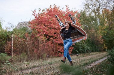 Güz parkı ormanlarında koşan sarışın güzel bir kadının uzun bir portresi. Genç kız, kot pantolon ve renkli hırka giyiyor, doğal arka planda poz veriyor, eğleniyor. Hippi tarzı..