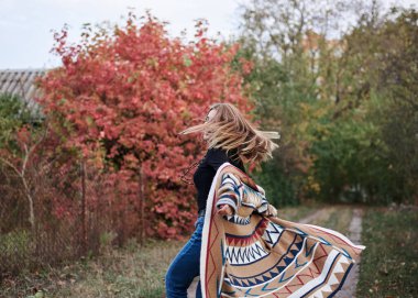 Güz parkı ormanlarında koşan güzel sarışın bir kadının çeyrek portresi. Genç kız, kot pantolon ve renkli hırka giyiyor, doğal arka planda poz veriyor, eğleniyor. Hippi tarzı..