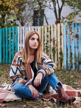 Genç sarışın kadın, renkli hırka ve mavi kot pantolon giyiyor, kırsalda renkli eski ahşap çitlerin yanında battaniyenin üzerinde oturuyor. Sonbaharda parkta poz veren hippi müzisyen..