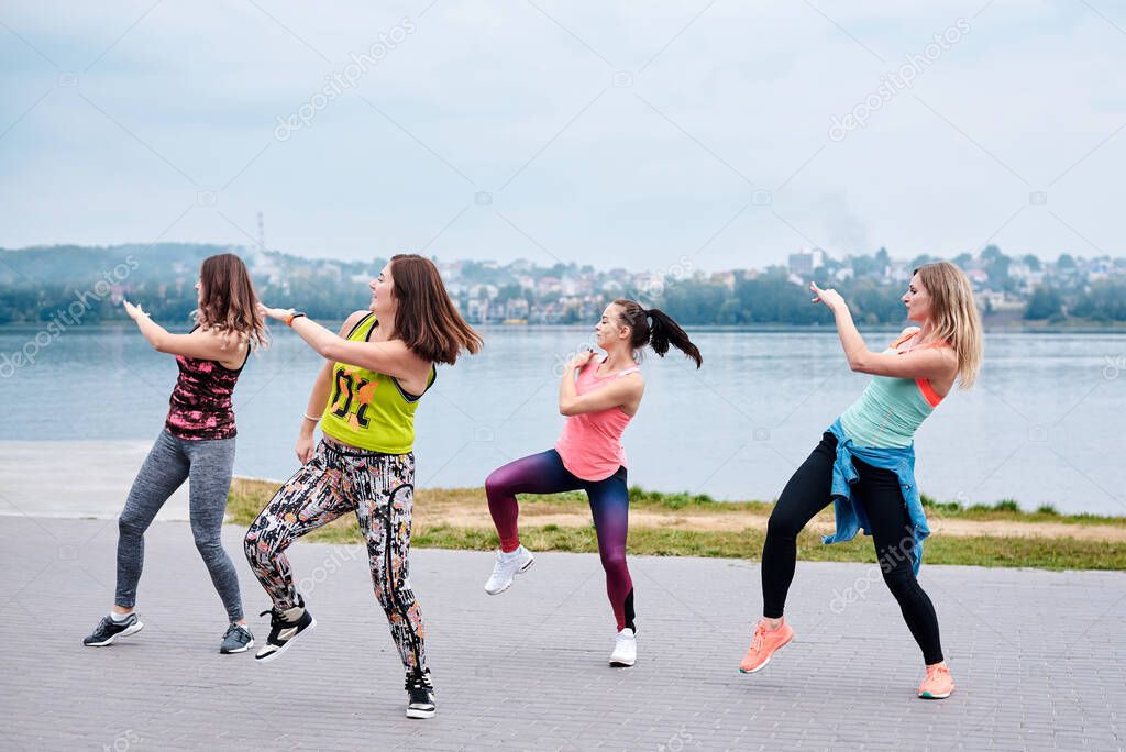 A group of young women, wearing colorful sports outfits, doing zumba exercises outside by city lake. Latin dance training to lose weight in summer. Healthy lifestyle concept. Female sport leisure.