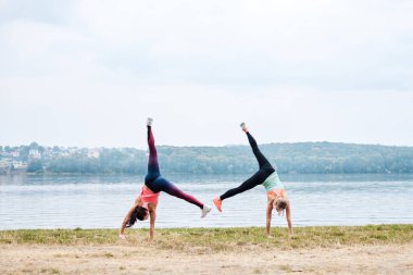 Renkli spor kıyafetleri giyen iki genç ve güçlü sporcu yazın şehir gölü kenarında çember egzersizi yapıyor. Sokak çalışması. Sağlıklı yaşam tarzı kavramı. Kadın güç eğitimi..