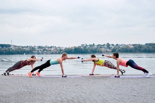 A group of four young women, wearing colorful sports outfit,doing fitness exercises on yoga mats outside by city lake in summer. Workout power female training to loose weight and body shape at nature.