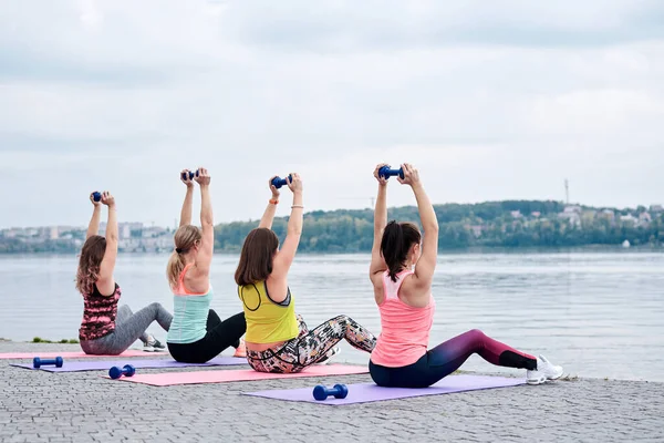 A group of four young women, wearing colorful sports outfit,doing fitness exercises on yoga mats outside by city lake in summer. Workout power female training to loose weight and body shape at nature.