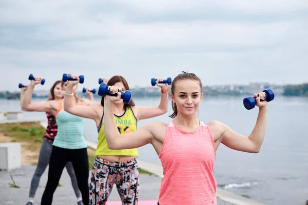 A group of four young women, wearing colorful sports outfit,doing fitness exercises with small weights outside by lake in summer.Workout power female training to loose weight and body shape at nature.
