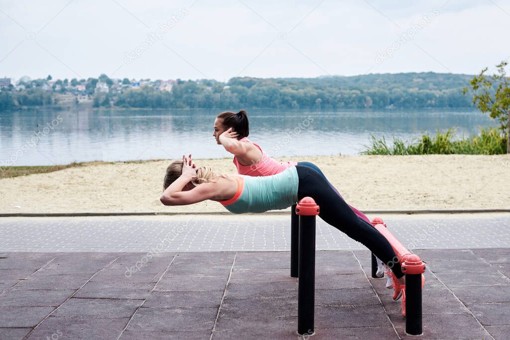 Two young women, wearing sports outfit, doing sit-ups on horizontal bars outside by city lake. Group of girls, fitness training together outdoors. Healthy lifestyle concept. Female athletic workout.