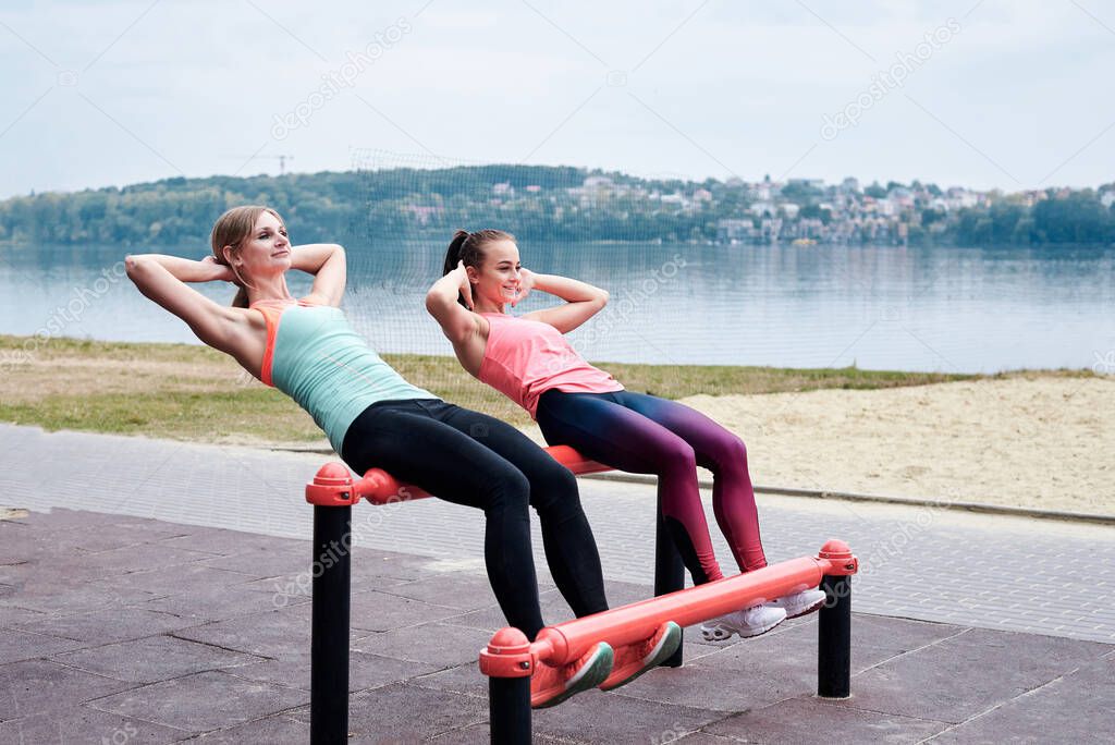Two young women, wearing sports outfit, doing sit-ups on horizontal bars outside by city lake. Group of girls, fitness training together outdoors. Healthy lifestyle concept. Female athletic workout.