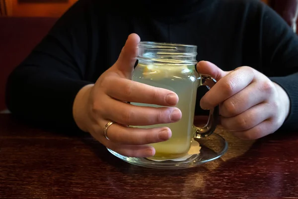 Vrouw die warme en verfrissende gemberthee drinkt met schijfje citroen in een restaurant. Handen vasthouden kopje natuurlijke kruidenthee op houten tafel, warme winterdrank — Stockfoto