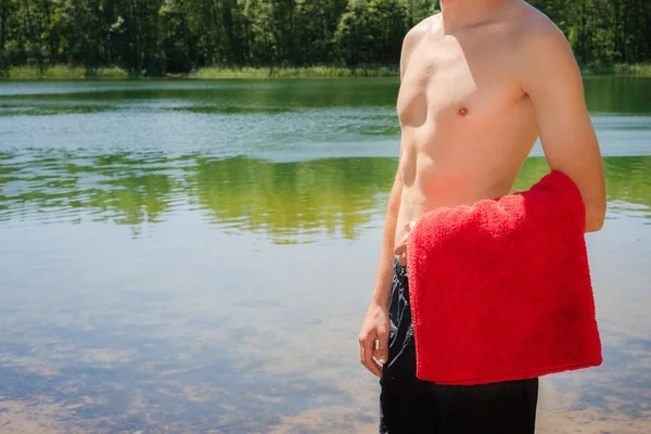 Ein junger, durchtrainierter, athletischer Mann steht ohne Hemd am Ufer des Sees und hält ein rotes Handtuch in der Hand. Schwimmen, Aktivitäten im Sommer in der Natur — Stockfoto