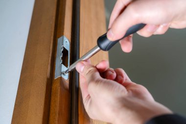 Carpenter repairing door lock. Installing a door handle. Handyman tightening door hinge . Hands of the repairman with a screwdriver. Locksmith screwing bolt into wooden door clipart