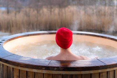 Genç yetişkin, dışarıda, tahta jakuzide dinleniyor ve doğayı seyrediyor. Güneşli bir günde sıcak havuzun keyfini çıkaran kişi, özel spa tedavisi
