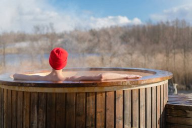 Genç yetişkin, dışarıda, tahta jakuzide dinleniyor ve doğayı seyrediyor. Güneşli bir günde sıcak havuzun keyfini çıkaran kişi, özel spa tedavisi
