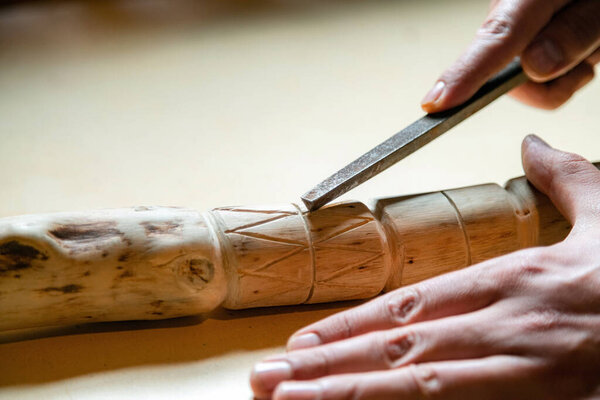 Process of man making wooden walking stick indoors during quarantine. Carving wood stick on the table using knife