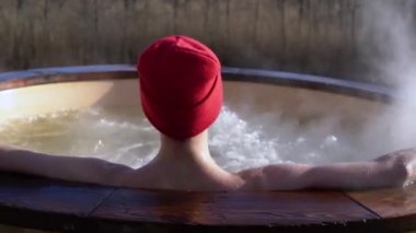 Genç yetişkin, dışarıda, tahta jakuzide dinleniyor ve doğayı seyrediyor. Güneşli bir günde sıcak havuzun keyfini çıkaran kişi, özel spa tedavisi