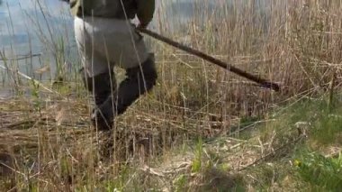 Lastik botları olan bir adam göl kıyısına doğru koşuyor. Güneşli bir günde yaşlı tırpanla zorbalık yapan bir adam.