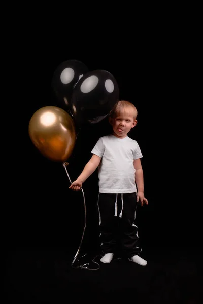 Blond batole s jazykem visí v bílém tričku drží černé a zlaté balónky na tmavém pozadí. — Stock fotografie