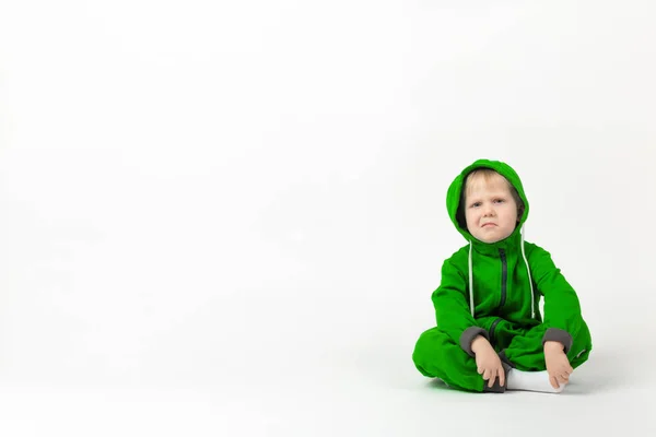 Un niño carismático disgustado con un mono verde brillante se sienta en el suelo y mira a la cámara con duda y desprecio . —  Fotos de Stock