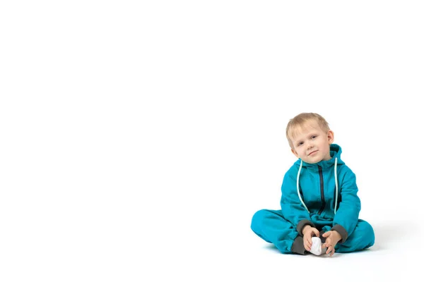 Malý blonďatý chlapec sedí na podlaze v tyrkysové kombinéze a usmívá se na kameru na bílém pozadí s místem pro text. — Stock fotografie