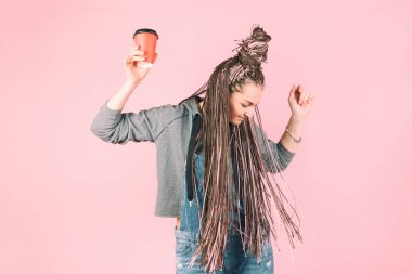 Pembe zizi afro örgülü, elinde bir fincan kahveyle pembe arka planda dans eden modern, güzel bir kız..