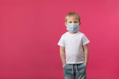 Sarışın Avrupalı bir çocuk Coronavirus 'un tıbbi maskesi içinde kameraya bakıyor. Pembe arka planda salgın konuları, korunma ve güvenlik için metin içeren bir stüdyo çekimi.
