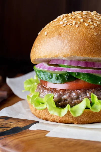 Burger on wood board at dark background. Sesame bun with beef, lettuce, tomato, cucumber and onion — Stock Photo, Image