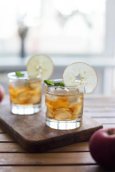 Apple juice with ice, mint and apple slice as garnish on wood board with light rustic background. Concept of seasonal beverage