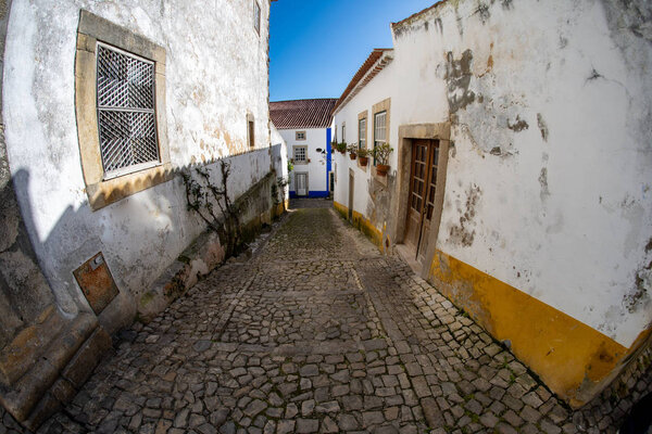 Fisheye lens view looking down a narrow cobblestone alley at brightly colored homes in Obidos Portugal
