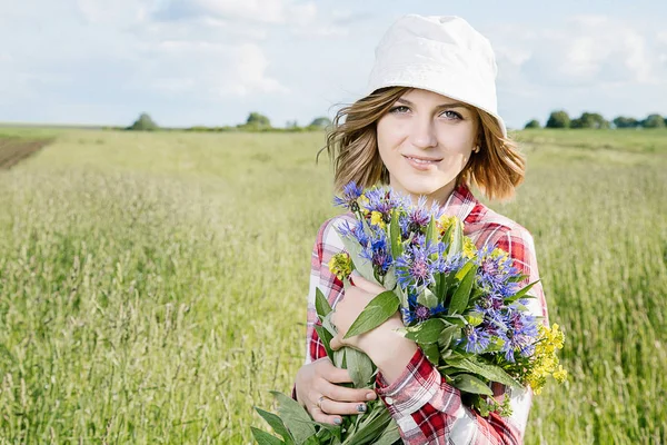 A girl in the field collects flowers, field flowers in a basket, a young girl with a smile, dressed in a shirt in a red box, in denim shirts and in a cappella, he goes and breaks colored (blue and yellow) flowers in a green field (meadow).