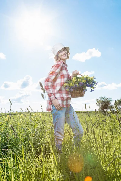 A girl in the field collects flowers, field flowers in a basket, a young girl with a smile, dressed in a shirt in a red box, in denim shirts and in a cappella, he goes and breaks colored (blue and yellow) flowers in a green field (meadow).