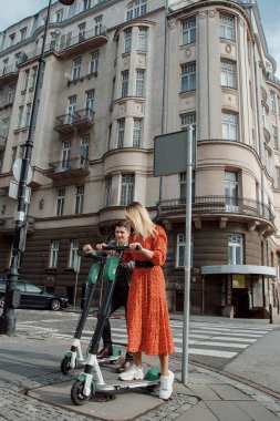 Genç ve sevgi dolu çift elektrikli scooterlarıyla modern cam bir binanın yanında oturuyorlar. Büyük şehirde yürür.