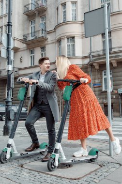 Genç ve sevgi dolu çift elektrikli scooterlarıyla modern cam bir binanın yanında oturuyorlar. Büyük şehirde yürür.