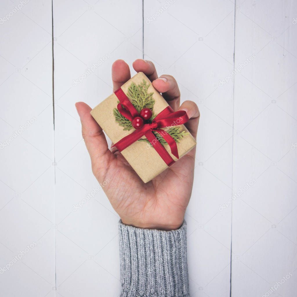 Woman's hand with gray sweater holding a little Christmas present on white wood background. Top view.