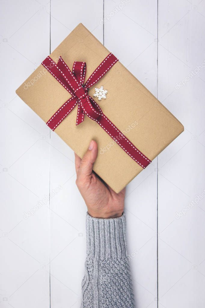 Woman's hand with gray sweater holding a Christmas present on white wood background. Top view.