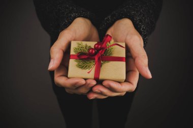 Elinde küçük bir Noel hediyesi tutan koyu renk kazaklı bir kadın. Siyah arkaplan.