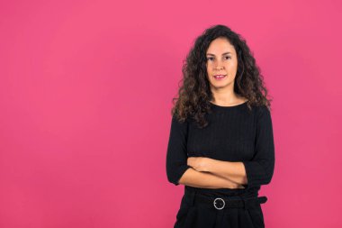 Kıvırcık saçlı esmer kadın ve siyah gömlekli kolları çapraz ve kameraya gülümseyen. Pembe arkaplan.
