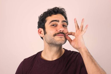 Kahverengi, gülümseyen, yakışıklı adam bıyığına mor tişörtle dokunuyor. Komik bir portre. Pembe arkaplan.